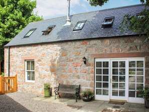 A stone building with a sloped roof, a wooden bench, and a door with multiple windows. - Sycamore Cottage - UK45923 (Ballater)
