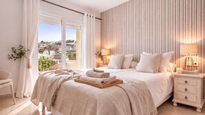 Un dormitorio con una cama grande, dos mesitas de noche, una silla y vistas a un paisaje urbano a través de una puerta corrediza.