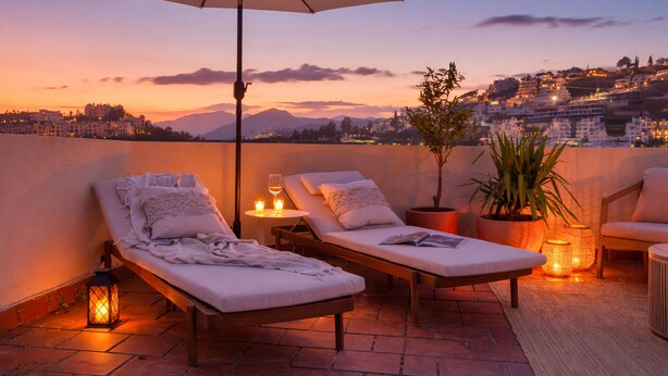 Una terraza en la azotea con sillones de descanso, una planta en maceta y vistas a un paisaje urbano al atardecer.