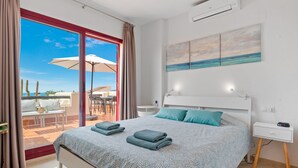 A hotel room with a bed, a nightstand, a desk, a chair, and a view of the sea through a glass door.