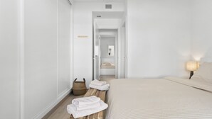 Een hotelkamer met een bed, een bedlamp, een houten bank met handdoeken, en een gang die naar een andere kamer leidt.