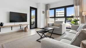 Een moderne woonkamer met een flatscreen televisie, een bank, een salontafel en grote ramen met uitzicht op gebouwen en groen.