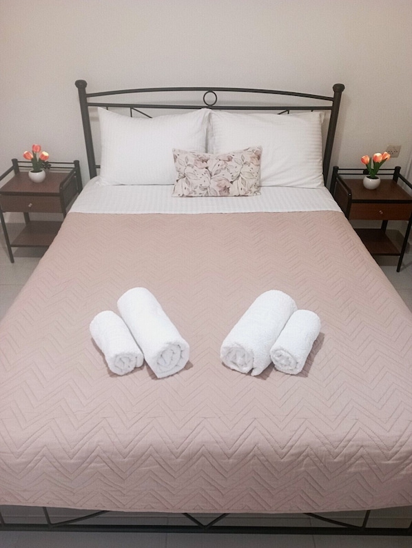 Un lit bien fait, avec deux serviettes enroulées, un coussin à motifs floraux et deux tables de chevet ornées de vases remplis de fleurs.