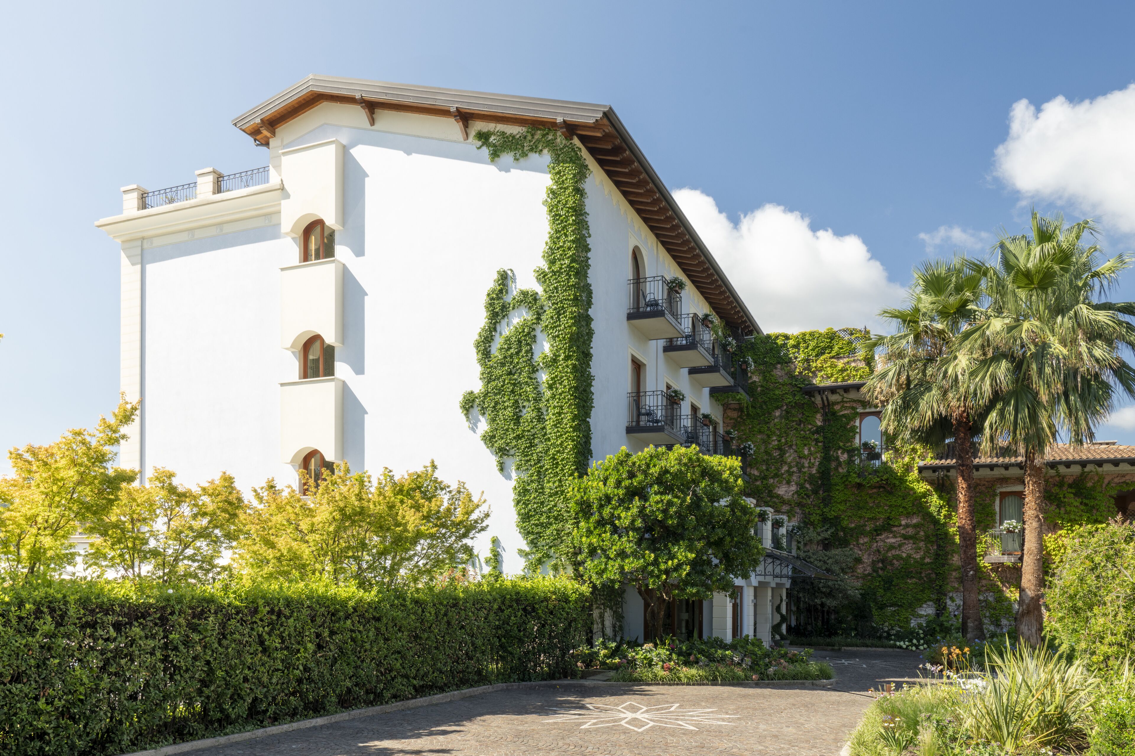 A white building with balconies, surrounded by greenery and palm trees.