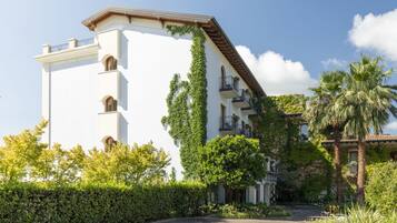 A white building with balconies, surrounded by greenery and palm trees.