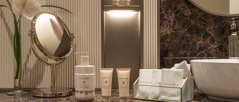 A bathroom vanity with a marble countertop, a round mirror, a white vase with a flower, and toiletries including a shoe brush and a shower cap.