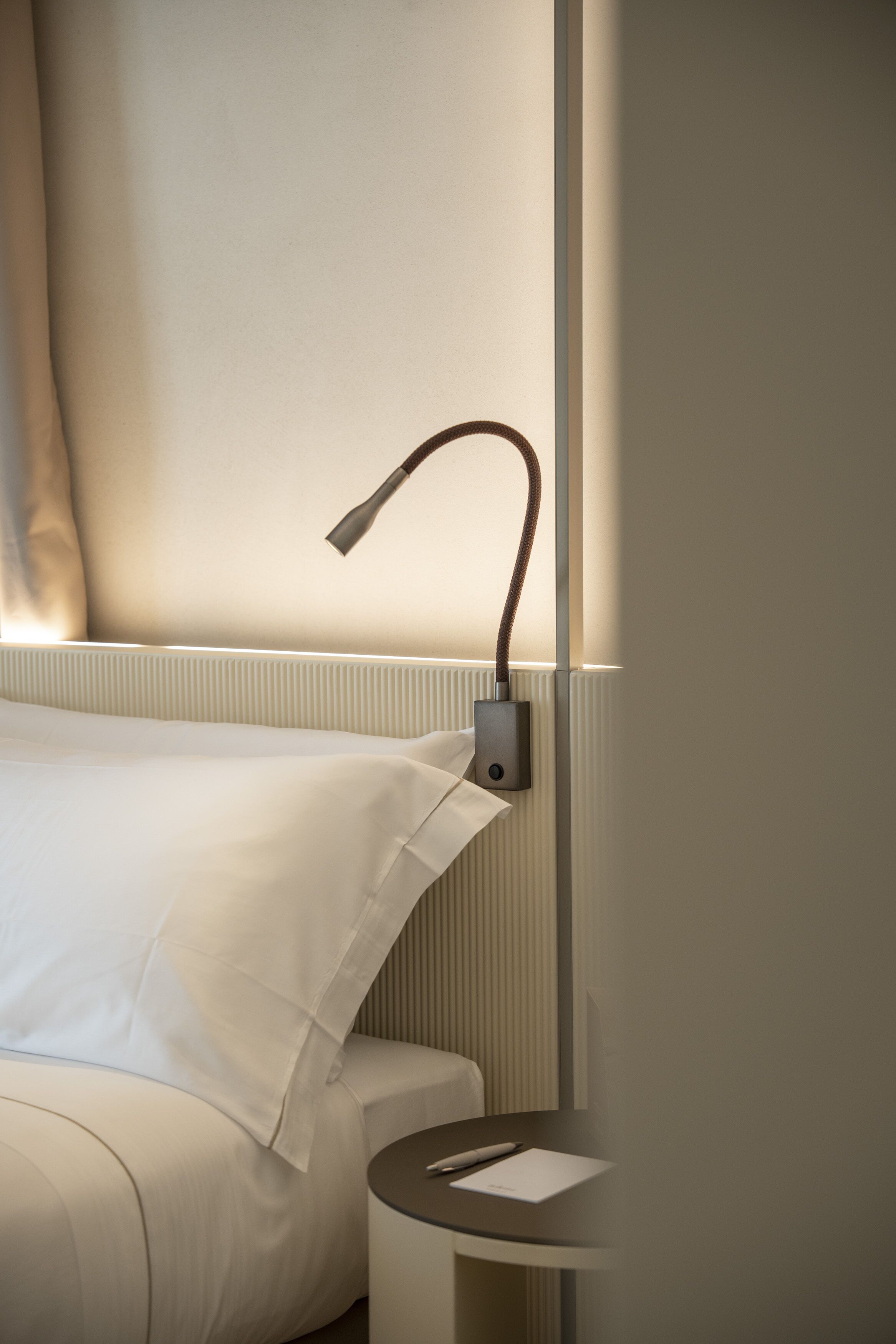 Un letto rifatto con lenzuola bianche, un comodino con una lampada e un punto luce a parete.