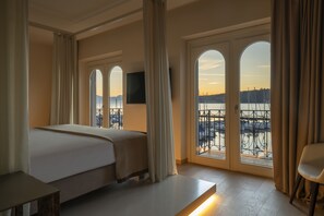 Habitación de hotel con cama, televisión y ventanas amplias que dan a una masa de agua y un paisaje urbano.