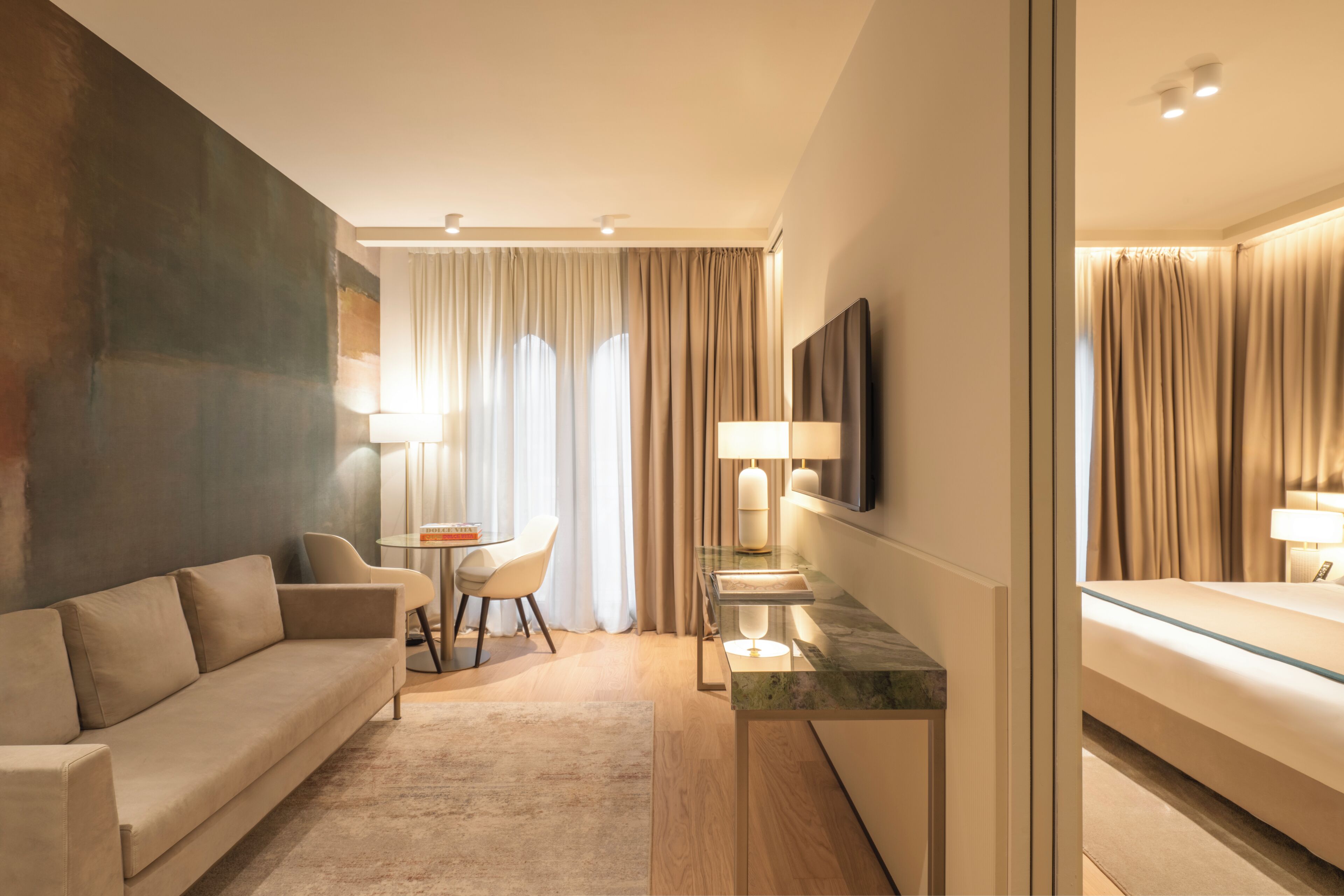 Une chambre d’hôtel moderne, équipée d’un canapé, d’une petite table, d’un téléviseur fixé au mur et d’un lit avec une tête de lit en verre.