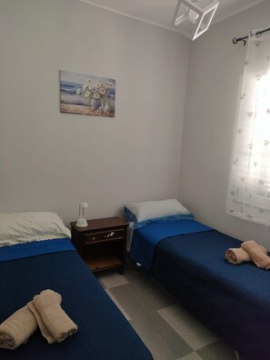 A small, simple room with two beds, a nightstand, a lamp, a painting on the wall, and a window with a curtain.
