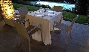 Ein Essbereich im Freien mit einem Tisch für vier Personen, umgeben von Korbstühlen, in der Nähe einer Steinsäule und einem Pool.