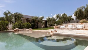 Una piscina amb un marc de pedra, una zona d'estar amb coixins blancs, i un edifici amb columnes al fons.