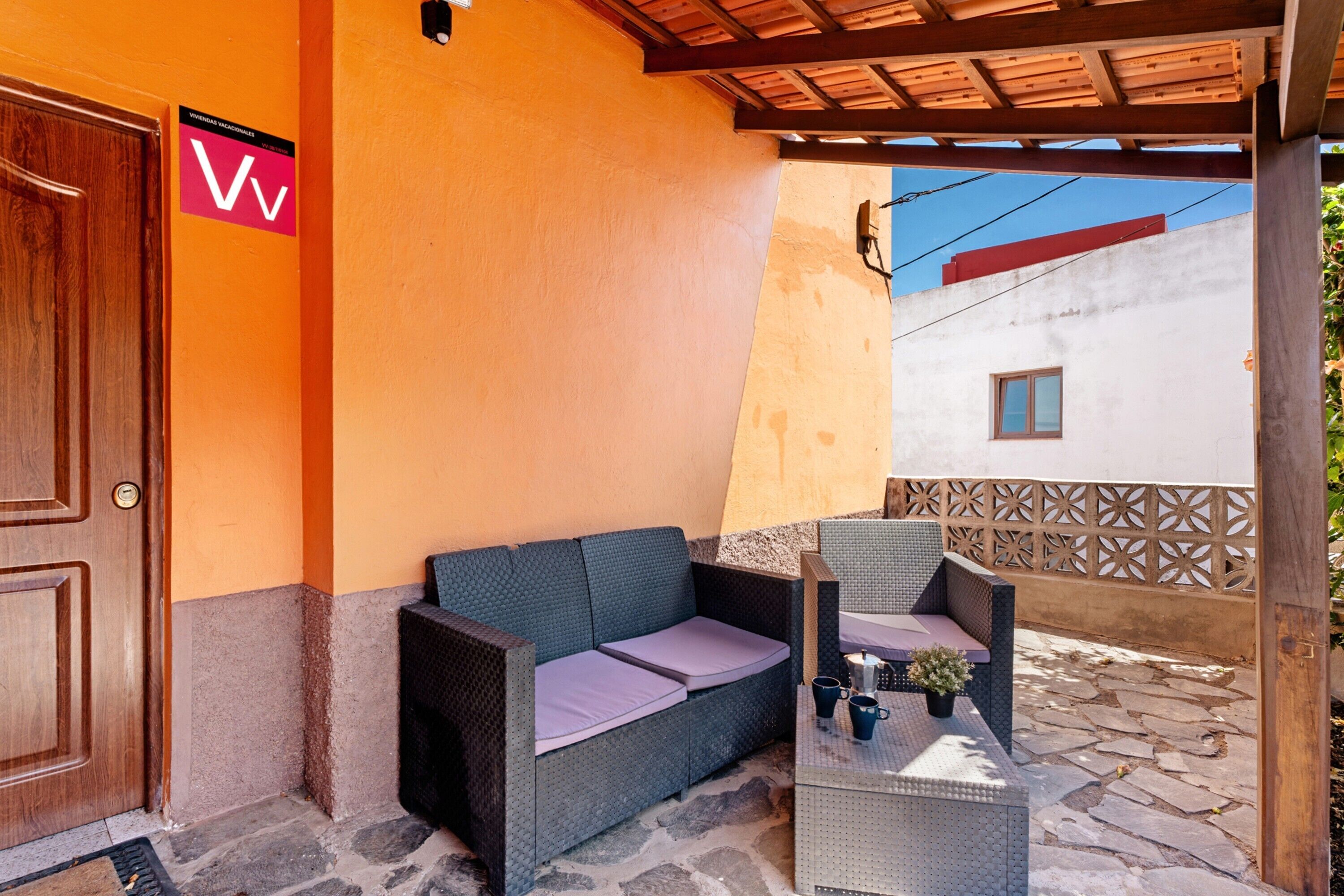 A covered outdoor seating area with a sofa and chairs, a door, and a wall with a sign.