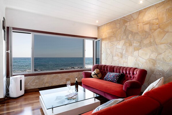 Une pièce avec un mur en pierre, un canapé rouge, une table basse en verre et une vue sur la mer.