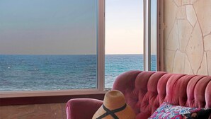 Une pièce avec une grande fenêtre donnant sur la mer, un canapé rouge et un chapeau tressé posé sur le canapé.