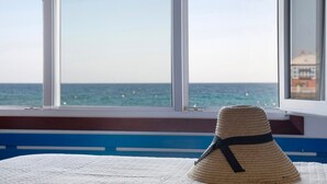Une chambre avec un lit, une fenêtre donnant sur la mer et un chapeau de paille posé sur le lit.