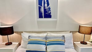 A neatly made bed with striped pillows, two bedside lamps, and a window with a cityscape view. - Groovy & Cool~1Bed/1Bath/1Car~Close to everything! (Bowen Hills)