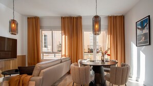 Een moderne woonkamer met een bank, een ronde eettafel en stoelen, grote ramen met gordijnen en hangende hanglampen.