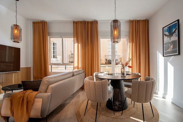 A modern living room with a sofa, round dining table, and chairs, large windows with curtains, and hanging pendant lights.