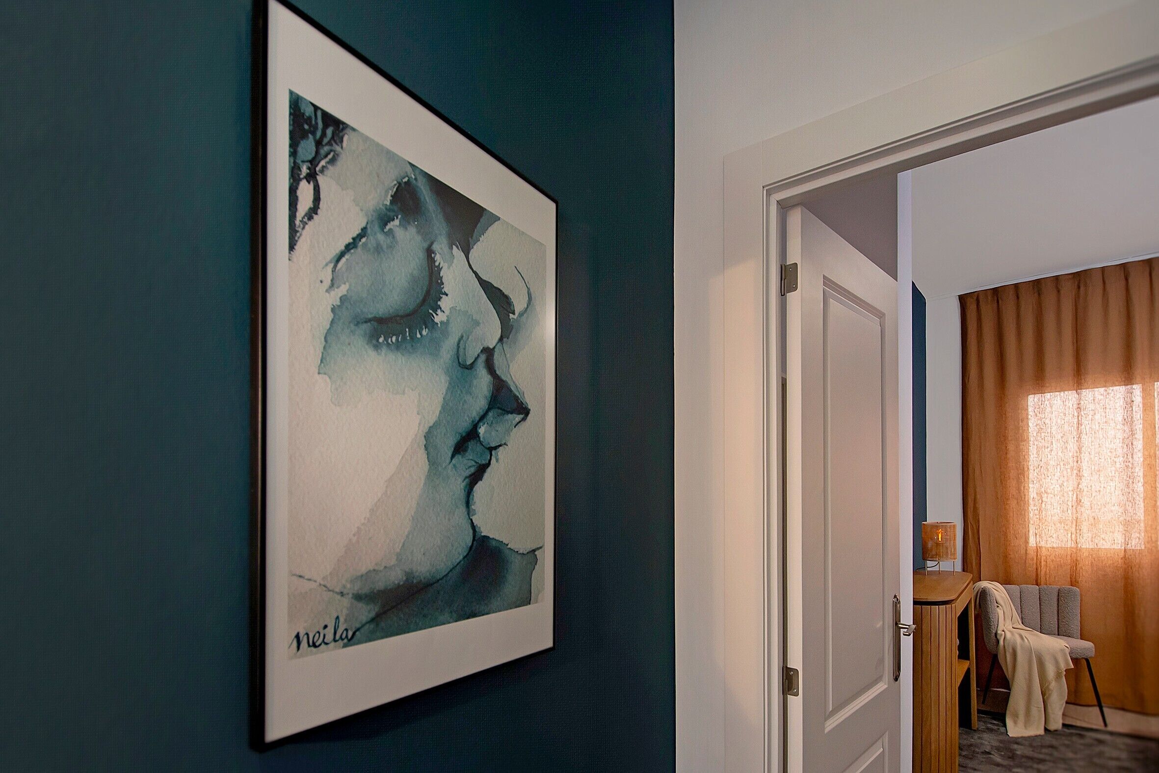 Una obra de arte enmarcada en una pared, una puerta que da a una habitación con una lámpara y una silla, y una ventana con cortinas.