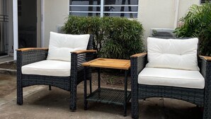 Two wicker chairs with white cushions and a small wooden table in an outdoor area with a concrete floor.