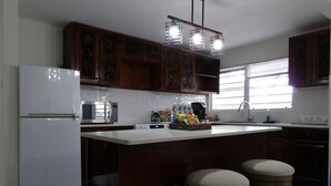 A kitchen with a white refrigerator, a microwave, a coffee maker, and a sink. There are two stools and a window with blinds.