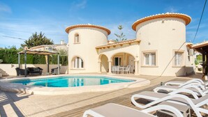 Un área de piscina con sillones, una pérgola y un edificio con ventanas arqueadas y techo de tejas.