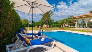 A swimming pool with lounge chairs and umbrellas, surrounded by greenery and a building with large windows.