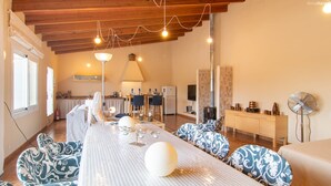 A spacious room with a long dining table, patterned chairs, a kitchen area, and a fireplace.