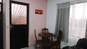 Ein Zimmer mit einer schwarzen Tür, einem Esstisch mit Stühlen, einer Couch, einem Fenster mit Vorhängen und einem Sonnenuntergangsbild an der Wand.