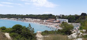 Escena costera con una playa, un edificio grande, sombrillas y gente.