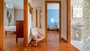 A room with a bed, a chair with a pink pillow, a mirror, and a bathroom with a sink and mirror.