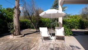 An outdoor patio with two white lounge chairs and a white umbrella, surrounded by trees and a building.