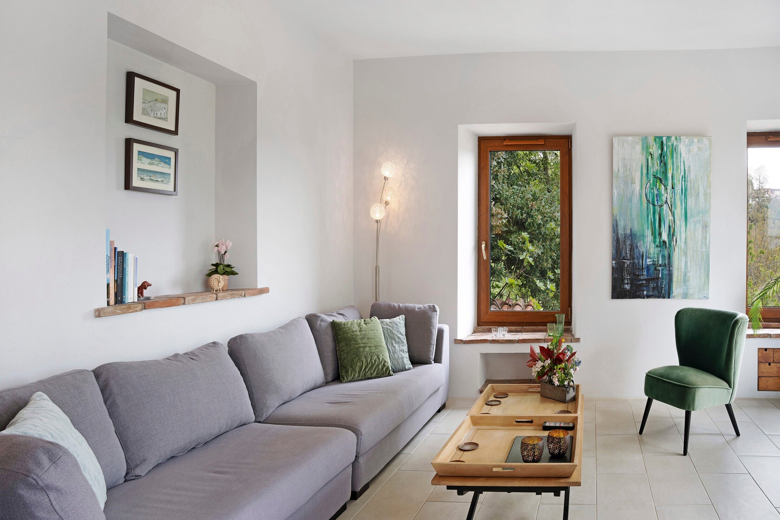 Un salon moderne avec un canapé gris, une table basse en bois et un fauteuil vert. Des tableaux encadrés ornent le mur, ainsi qu’une lampe sur pied et une fenêtre donnant sur un paysage verdoyant.