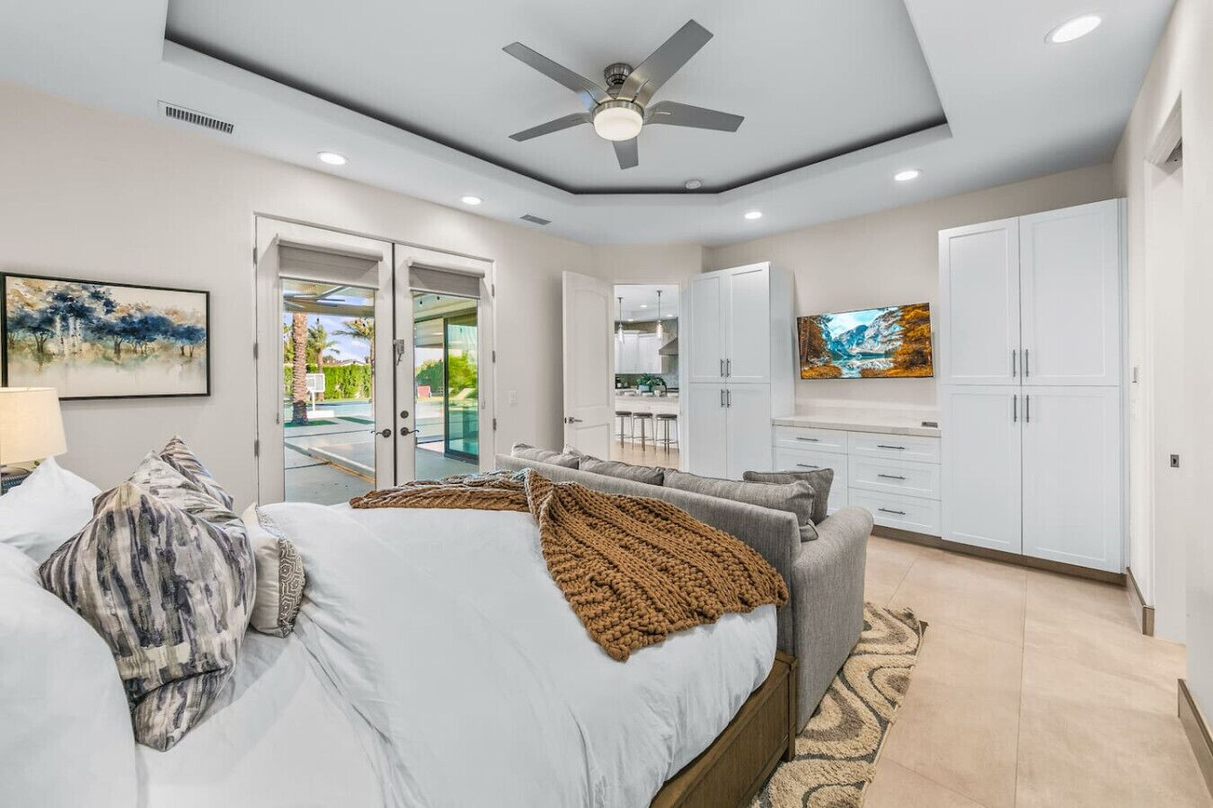 A modern bedroom with a bed, a ceiling fan, and a view of the kitchen through an open door.