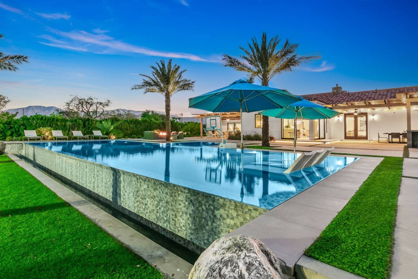 A rectangular swimming pool with a clear water surface, surrounded by a well-maintained lawn and a patio area with outdoor seating.