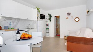 Una cocina moderna con armarios blancos, una mesa de comedor con una botella de vino y naranjas, un sofá y un televisor sobre la encimera de la cocina.