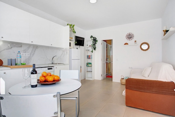 A modern kitchen with white cabinets, a dining table with a bottle of wine and oranges, a sofa, and a TV mounted above the kitchen counter.