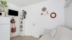 Un espacio habitable pequeño con kitchenette, televisor de pantalla plana, sofá blanco y una puerta que da a otra habitación.