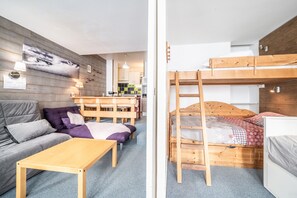 Une pièce comprenant un lit superposé, un canapé, une table, un coin cuisine et un mur orné d’un tableau.