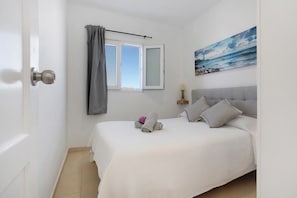 A bedroom with a bed, a window with curtains, a painting, and a bedside table with a lamp.
