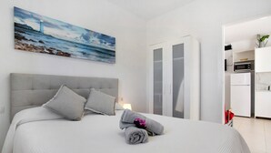 Uma cama bem arrumada com travesseiros, um quadro de um farol e uma cozinha visível através de uma porta aberta.
