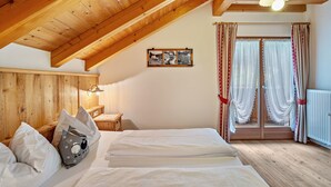 Een slaapkamer met een schuin houten plafond, een bed met wit beddengoed, een nachtkastje met een lamp en een raam met gordijnen.