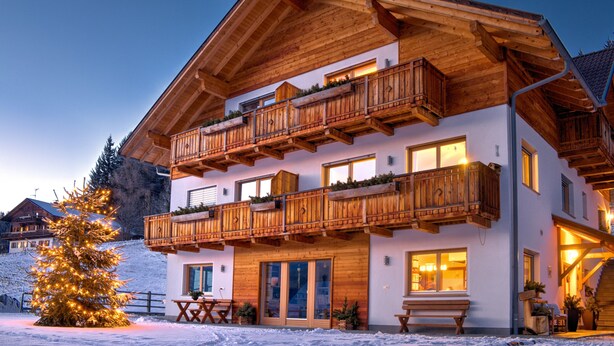 Een houten chalet met twee verdiepingen, voorzien van balkons, een houten terras en een sneeuwbedekte grond.