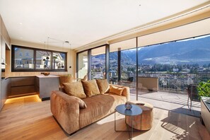 A modern living room with a sofa, a coffee table, and a view of a cityscape through large windows. - Holiday Apartment 'Levita Living Wolke' with Mountain View and Private Terrace (Dorf Tirol)