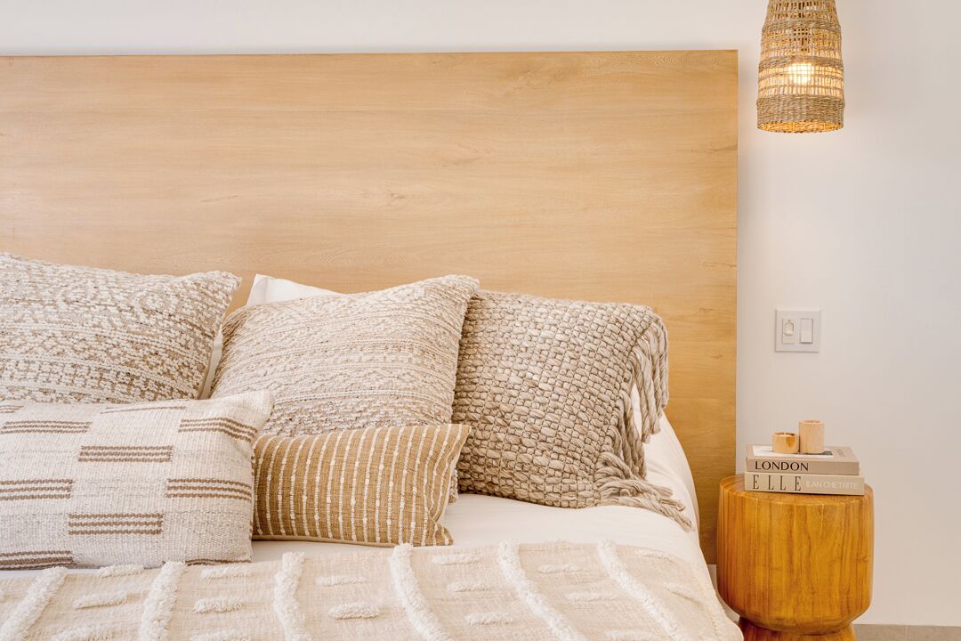 Un lit bien fait, agrémenté de plusieurs oreillers et d’une couverture, une tête de lit en bois, une petite table de chevet en bois avec des livres, et une suspension lumineuse en fibres tressées.