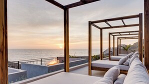 Eine Dachterrasse mit Meerblick bei Sonnenuntergang, ausgestattet mit einer Holz-Pergola, weißen Sitzgelegenheiten mit Kissen und einer Feuerstelle.