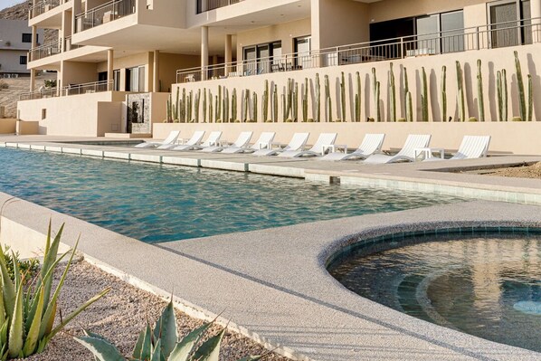 A pool area with lounge chairs, a cactus wall, and a building with balconies.