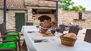 Un espace extérieur aménagé pour les repas, avec des murs en pierre, un four en briques, des chaises en osier et une table dressée pour deux personnes, avec des verres à vin et du pain.