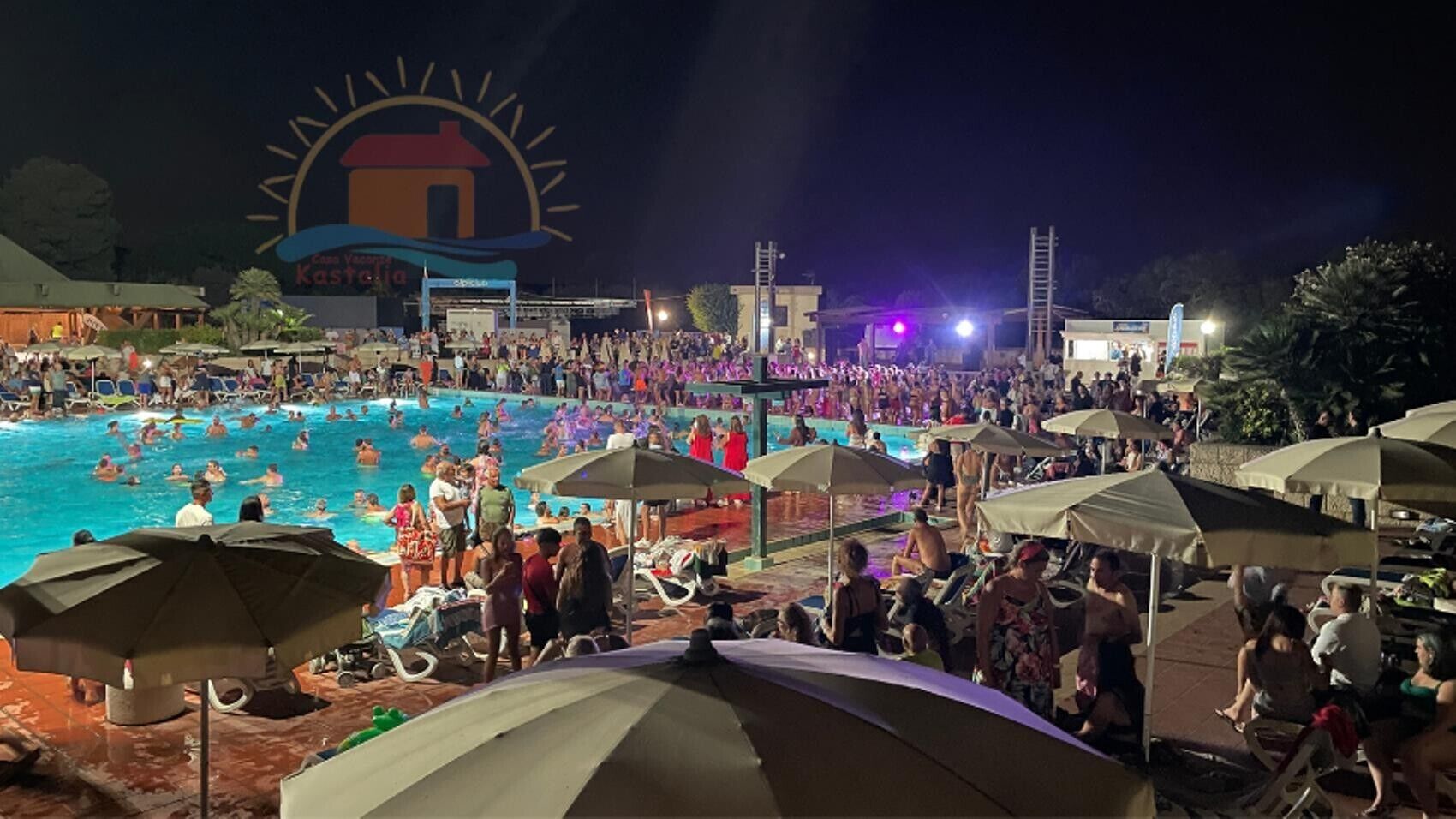 Une grande piscine extérieure où se trouvent de nombreuses personnes, des parasols et, au fond, une scène.
